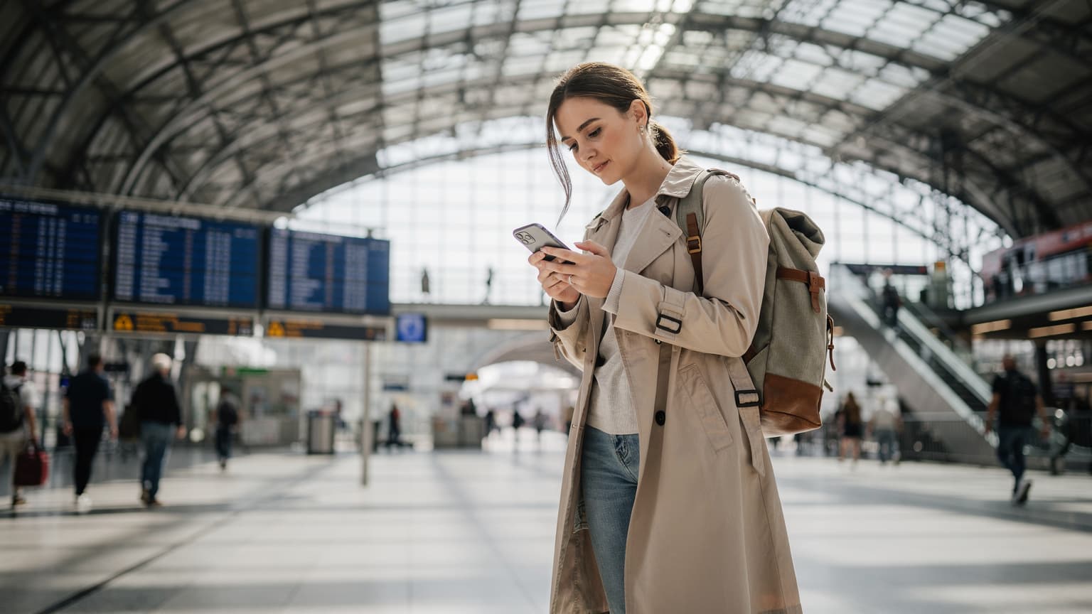 A woman confidently navigating a bustling train station in Berlin using her smartphone, with digital train schedules visible in the background.