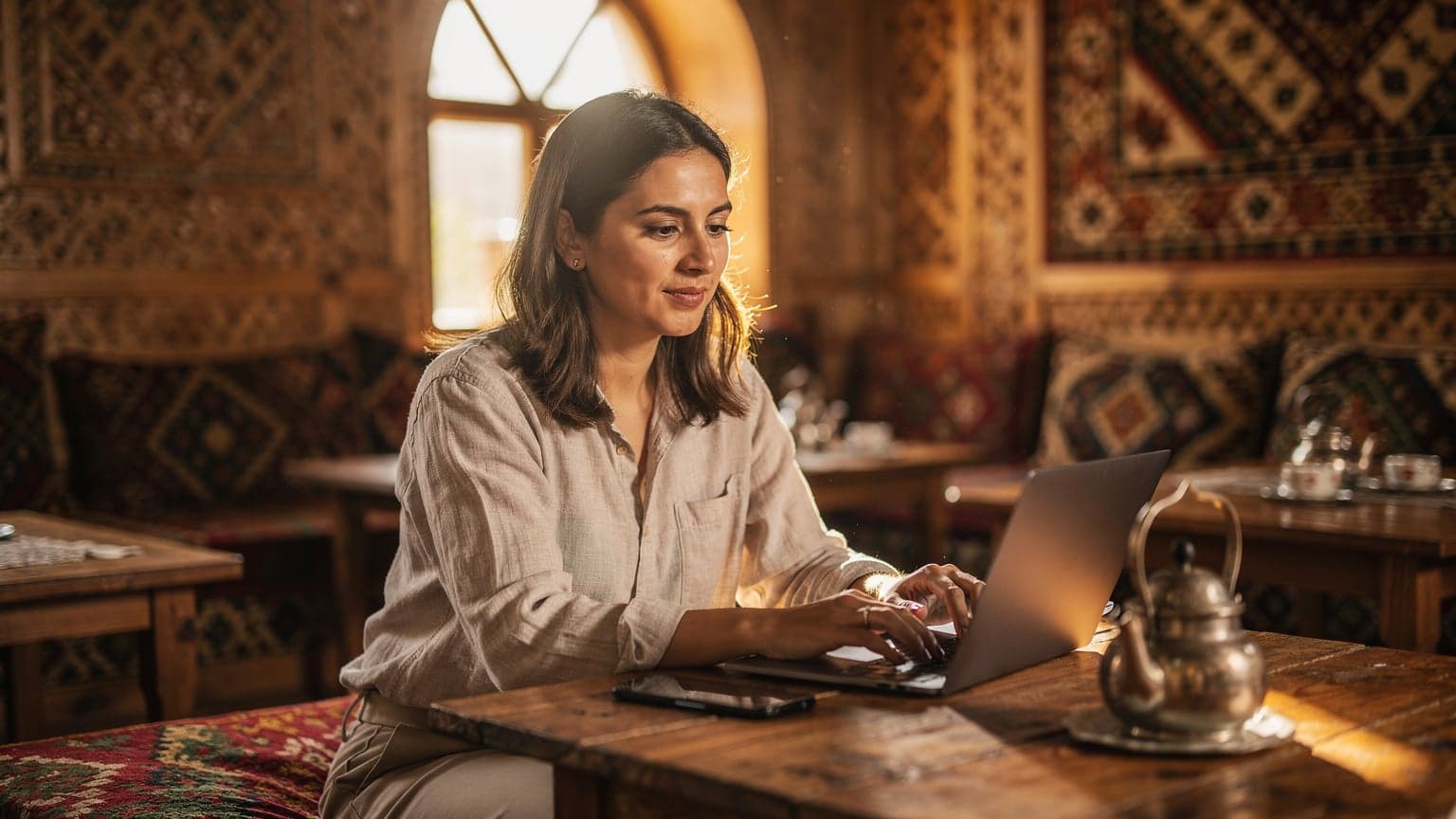 A digital nomad sits at a wooden table in a traditional Central Asian tea house in Tashkent, Uzbekistan, working on a laptop with a Cellesim eSIM enabled phone beside them, illuminated by soft morning light filtering through an arched window.