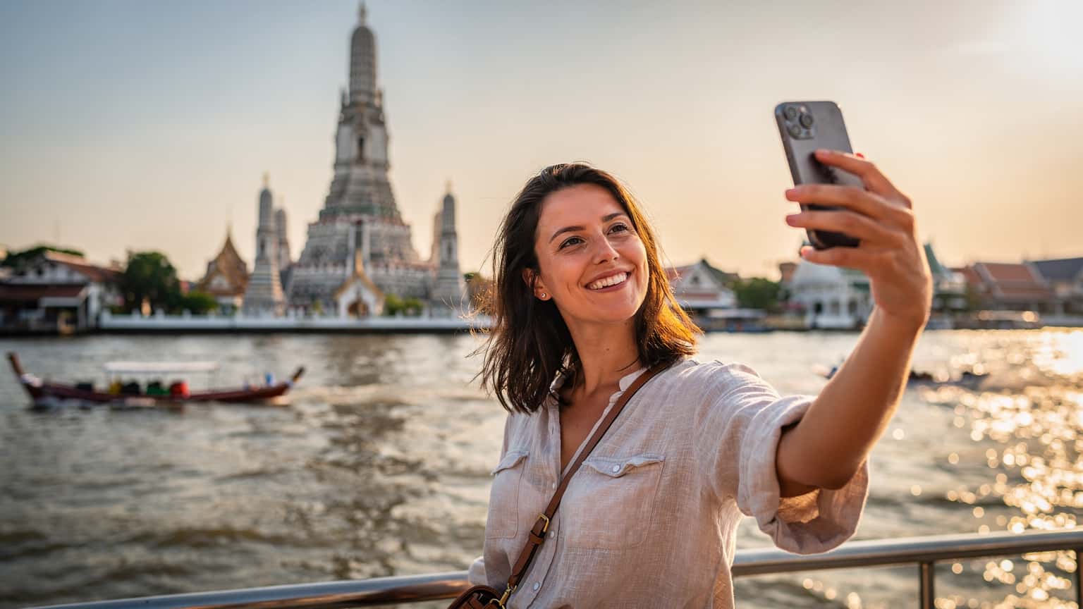 Une voyageuse souriante prend un selfie avec son téléphone devant le Wat Arun à Bangkok, au coucher du soleil, reflétant l'eau scintillante du fleuve Chao Phraya.