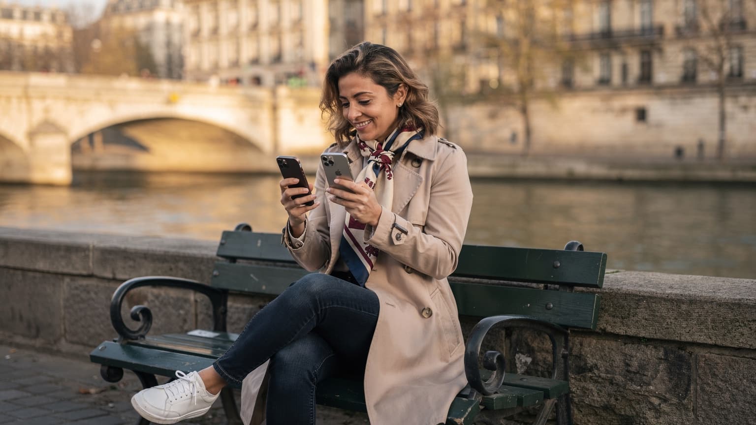 Une femme souriante, assise sur un banc public près de la Seine à Paris, tenant son nouvel iPhone et son ancien iPhone, comparant les écrans pour un transfert d'eSIM réussi, avec le Pont Neuf en arrière-plan.