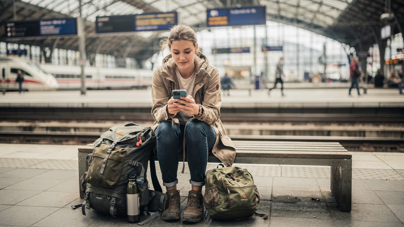 Eine junge Frau mit Rucksack sitzt auf einer Bank am Bahnhof München, ihr Smartphone in der Hand, auf dem der Cellesim-Tarif ausgewählt ist.