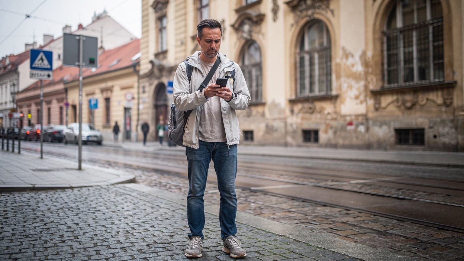 Muž kontroluje telefon v ruce před historickou budovou v Praze, zatímco po ulici projíždí tramvaj.
