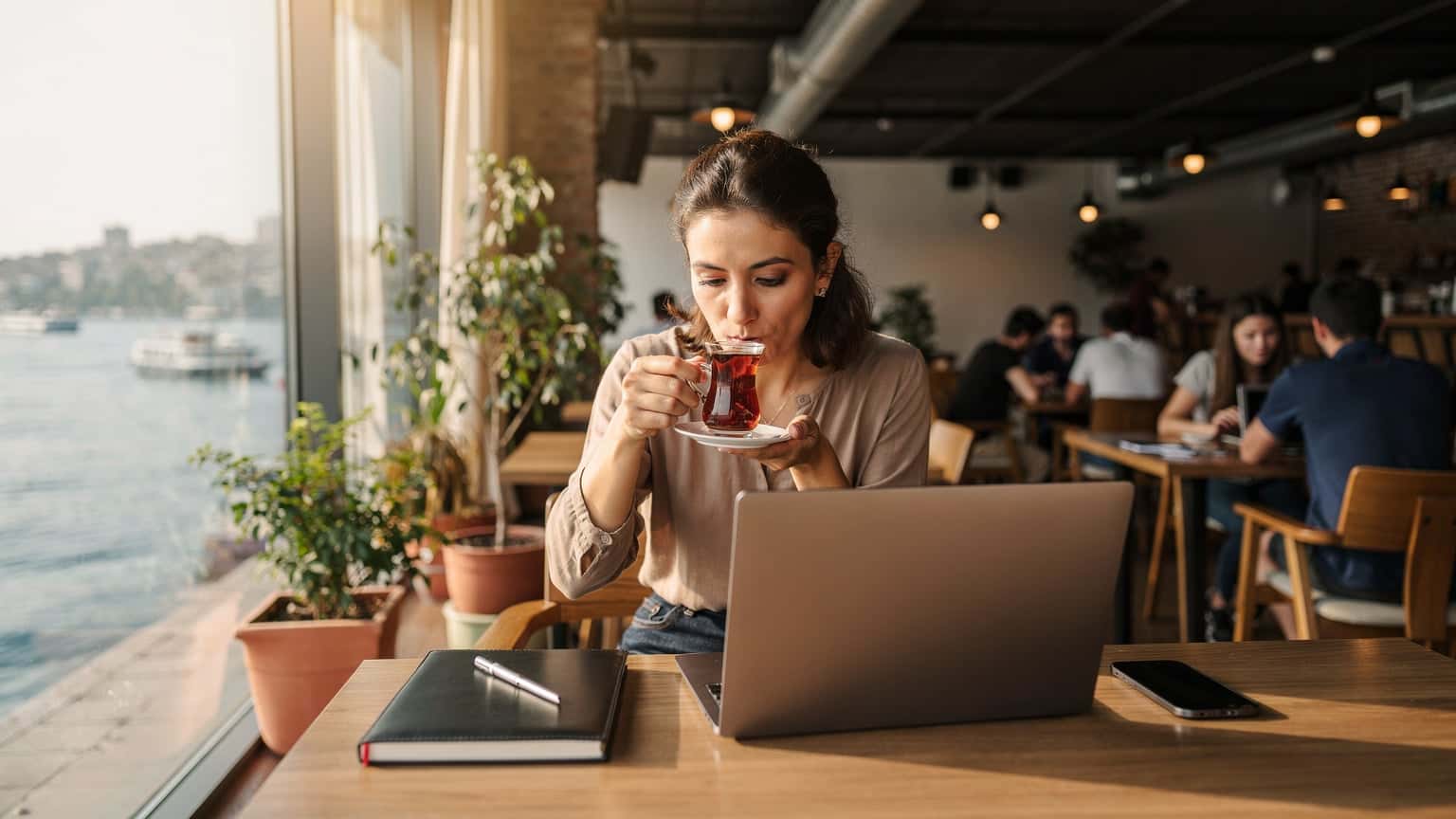 Eine Frau sitzt in einem Co-Working Space in Kadıköy, Istanbul, trinkt türkischen Tee und arbeitet an ihrem Laptop, während sie ihr Smartphone neben sich hat.