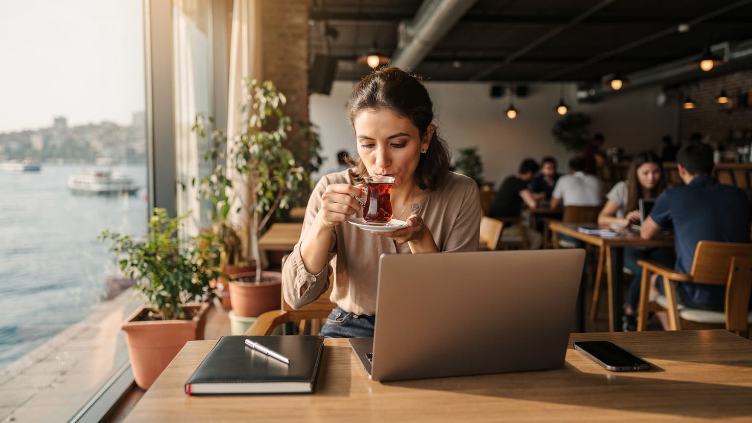 Eine Frau sitzt in einem Co-Working Space in Kadıköy, Istanbul, trinkt türkischen Tee und arbeitet an ihrem Laptop, während sie ihr Smartphone neben sich hat.