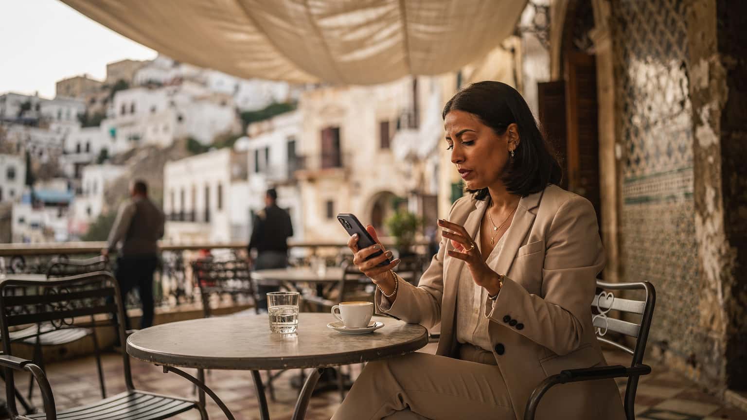 Une femme utilisant son smartphone avec une eSIM active, assise à la terrasse d'un café traditionnel à Alger, avec la Casbah en arrière-plan sous un ciel clair.