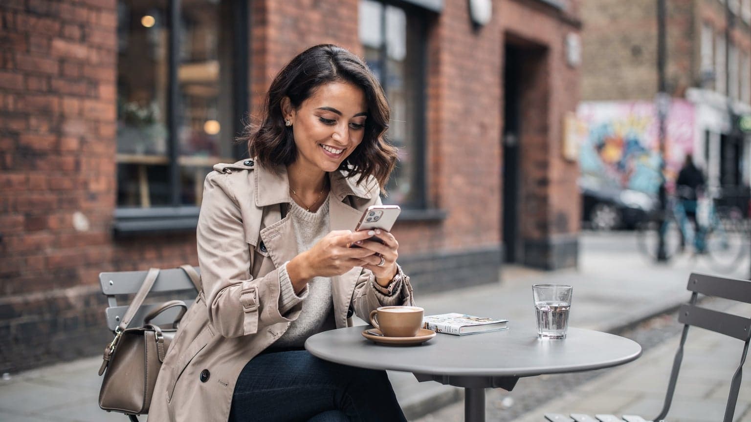 Una viaggiatrice italiana sorride mentre tiene il suo smartphone con un'eSIM attiva, seduta a un tavolo esterno di un caffè nel quartiere di Shoreditch a Londra, con un tipico edificio in mattoni rossi sullo sfondo.