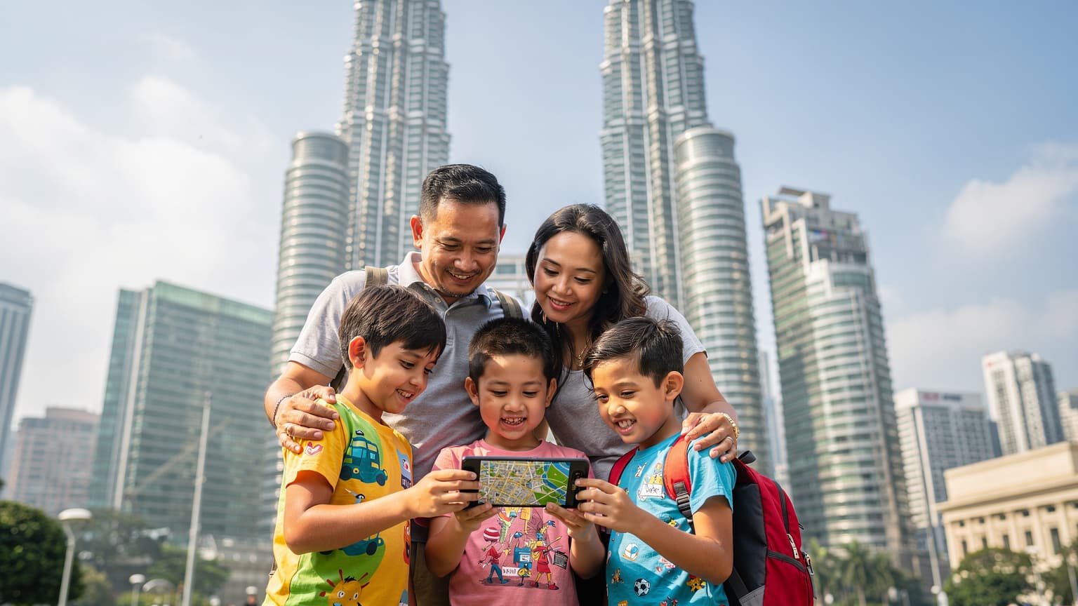 Sebuah keluarga Malaysia yang riang sedang melihat peta digital pada telefon pintar di hadapan Menara Berkembar Petronas, Kuala Lumpur, bersedia untuk perjalanan antarabangsa dengan beg bagasi mereka.