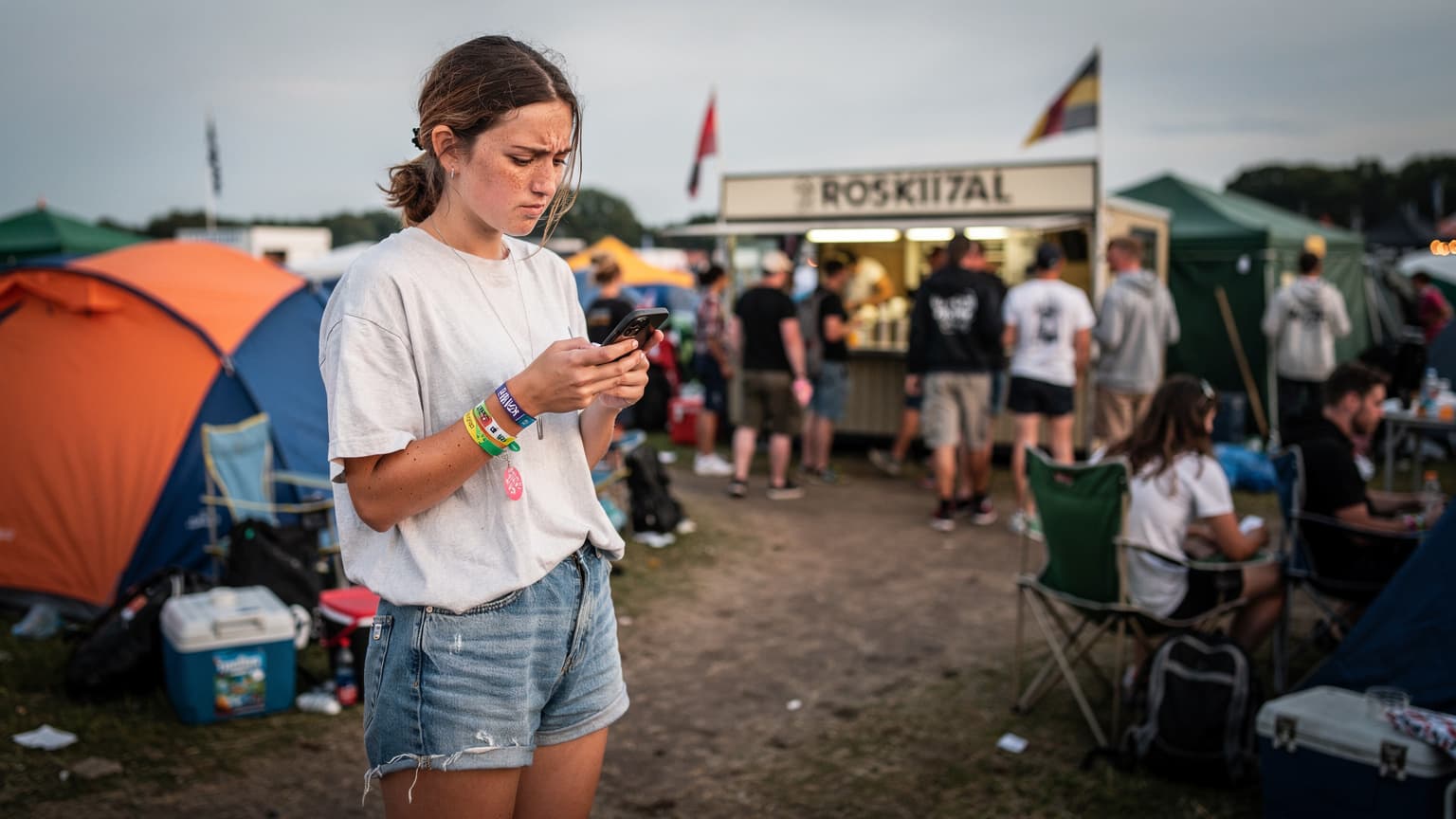 En ung kvinde med festivalarmbånd tjekker sin telefon på en travl festivalplads i Roskilde, omgivet af telte og glade mennesker.