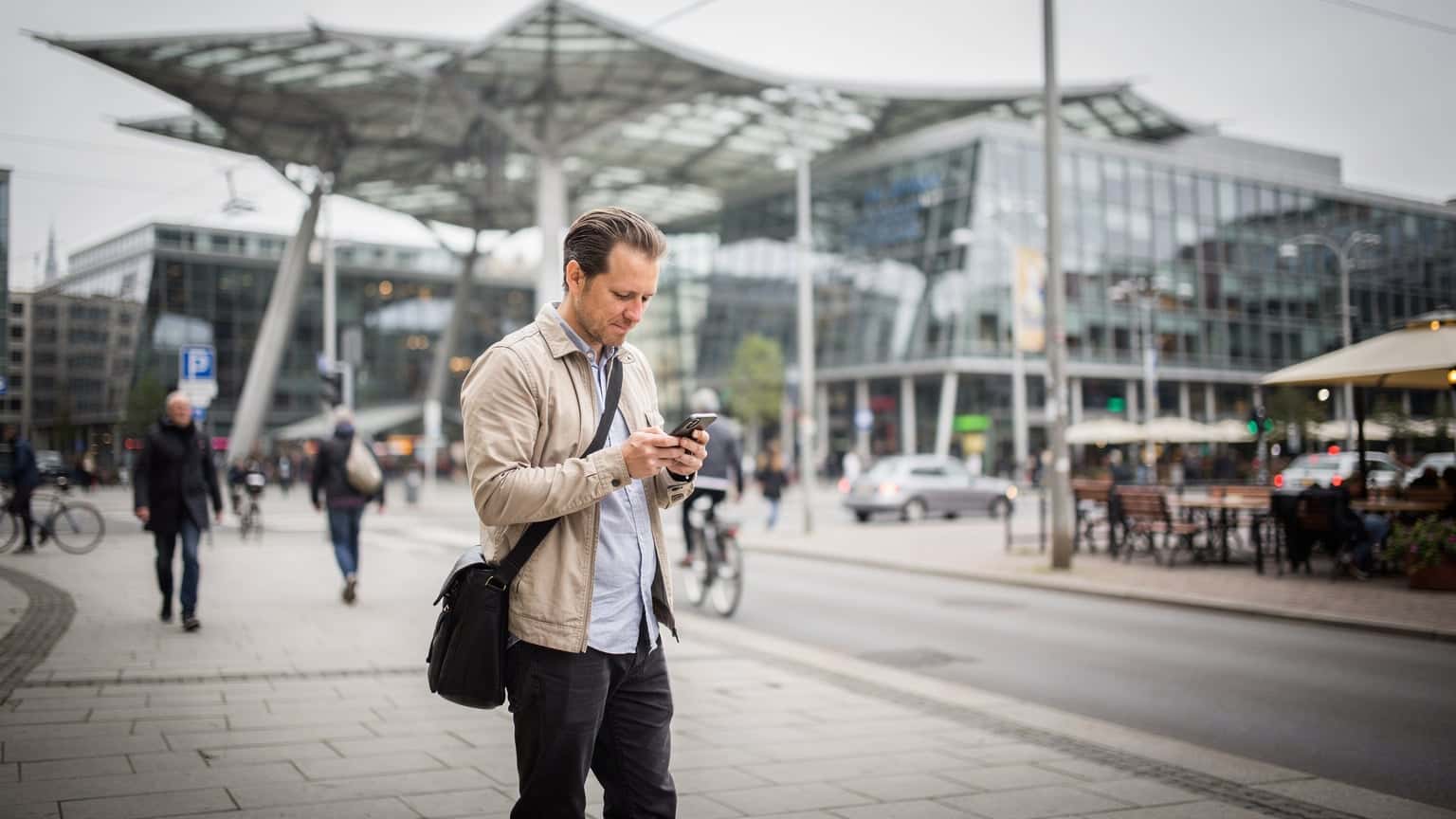 Un viaggiatore controlla il suo smartphone sul marciapiede della Potsdamer Platz a Berlino, con moderni edifici in vetro che riflettono il cielo, suggerendo connettività in un ambiente urbano.