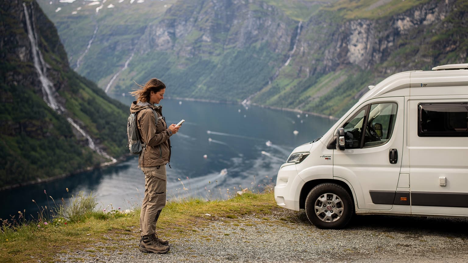 Kobieta stojąca przy kamperze na tle majestatycznego norweskiego fiordu, przeglądająca mapę na smartfonie z eSIM.