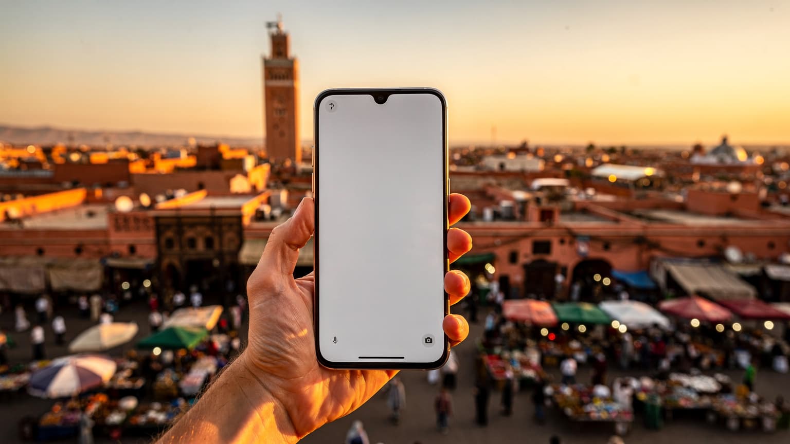 A modern smartphone is held in a hand, with the vibrant and bustling Jemaa el-Fna square and the Koutoubia Mosque in Marrakesh visible in the soft-focus background, illuminated by warm, natural golden hour light. The image depicts the integration of technology and travel.